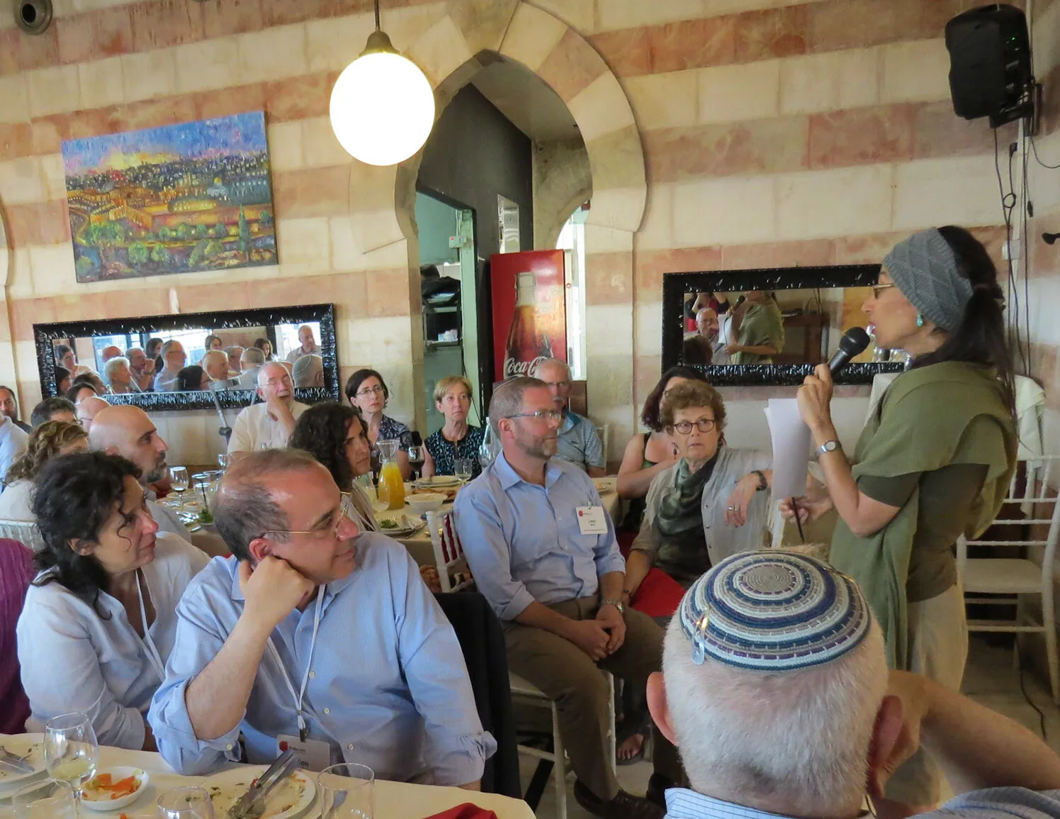 A woman speaking into a microphone to a seated audience at lunch tables in a stone-arched Jerusalem dining room