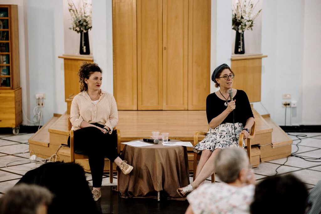 Two women in conversation on stage before an audience in the Pardes Beit Midrash