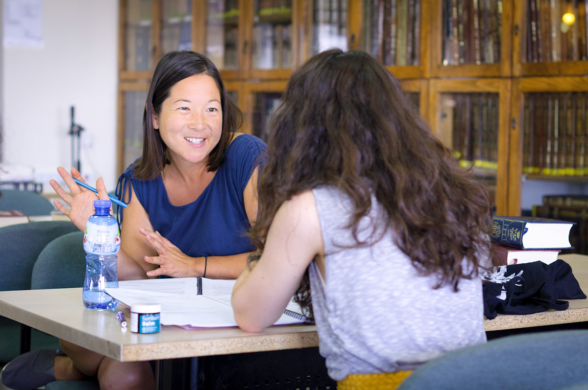 Two women in conversation across a table in the beit midrash, one gesturing as she explains a text