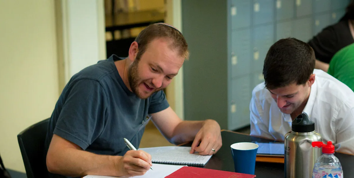 A smiling student writing notes in a spiral notebook at a table, with another student studying beside him