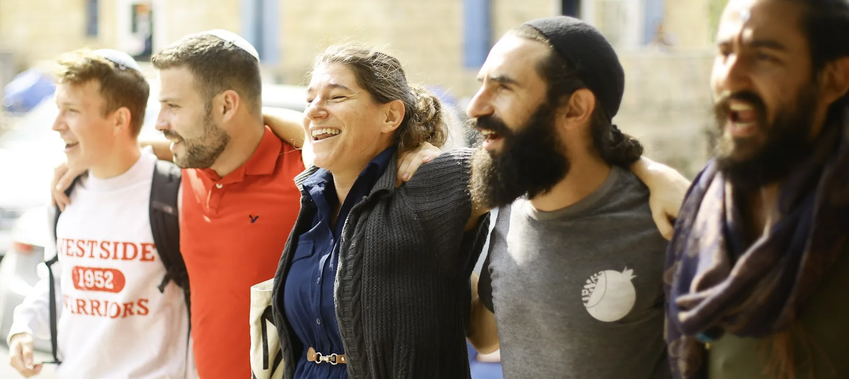 Five students standing with arms around each other outdoors, laughing and singing together