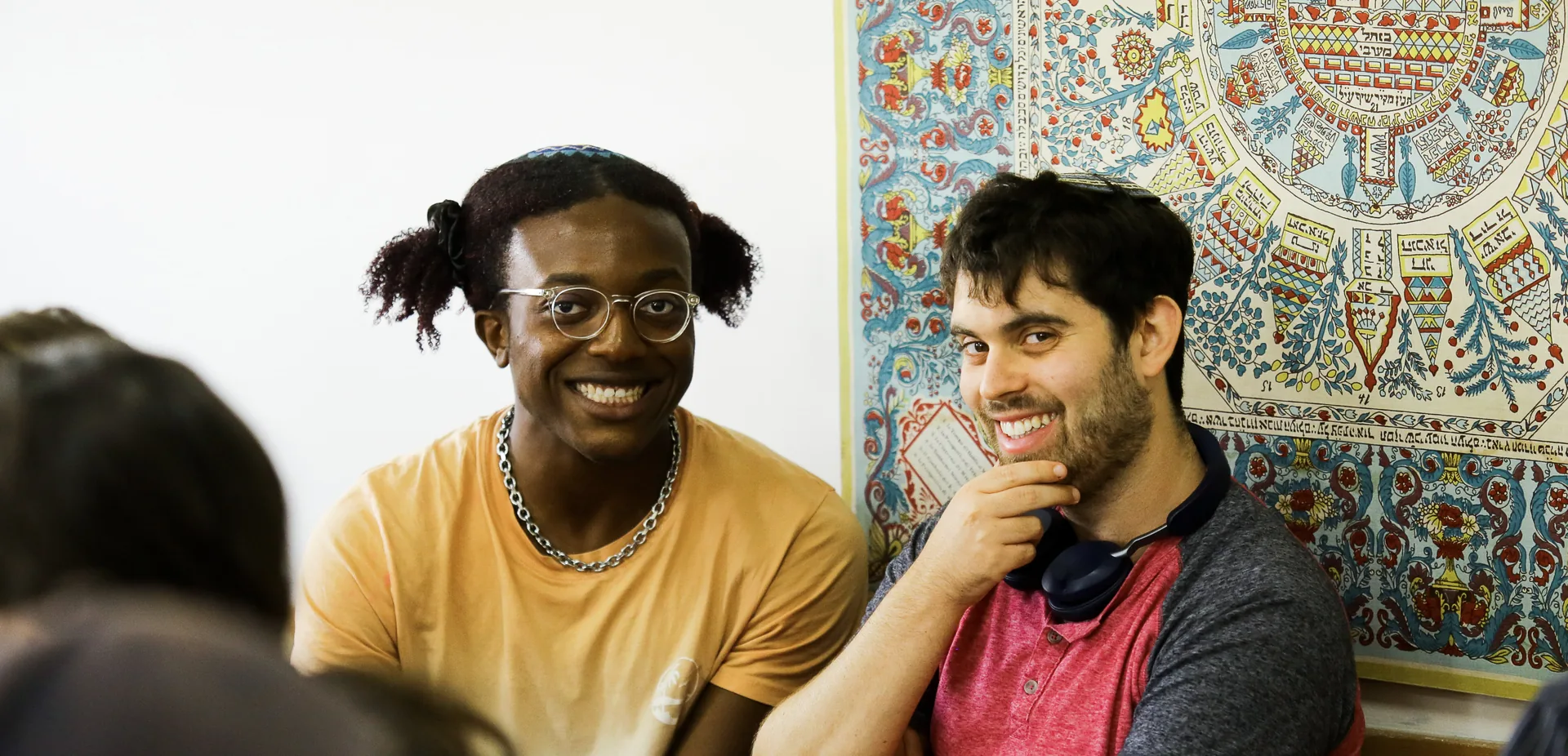 Two smiling students sitting in a classroom beside a decorated Hebrew manuscript poster, listening during a discussion